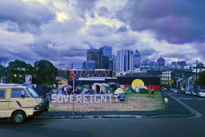 Image Description: The Block, the word sovereignty stands in big letters at front on vacant land where Aboriginal housing once stood. Now occupied by Aboriginal Elders camping in tents to protest commercial development plans. Iconic Aboriginal flag on the wall of the gymn in background. Sydney city rising up behind on the sky line. Lots of dark clouds overhead.