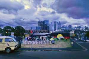 Image Description: The Block, the word sovereignty stands in big letters at front on vacant land where Aboriginal housing once stood. Now occupied by Aboriginal Elders camping in tents to protest commercial development plans. Iconic Aboriginal flag on the wall of the gymn in background. Sydney city rising up behind on the sky line. Lots of dark clouds overhead.