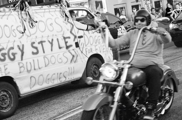 Black and white photograph of white van with 'dogs', 'doggies', 'doggy style' spray painted across the side, streamers hanging off it driving one direction and a biker driving the other direction. The street is full of fans, waving flags.