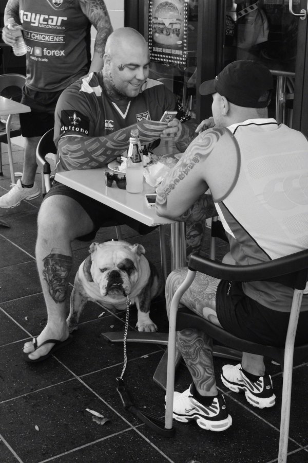 Black and white photograph of two men, Bulldogs fans, sitting at a table on the street in Belmore. A bulldog on a leash sits under the table.