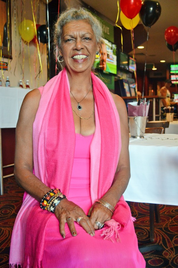 <strong>Image Description:</strong> Colour-photo portrait of Jenny Munro sitting on a chair. It's her 60th birthday celebration at the Abbotts Hotel, Waterloo. She's wearing a bright pink dress. There are yellow, red and black balloons floating in the background.