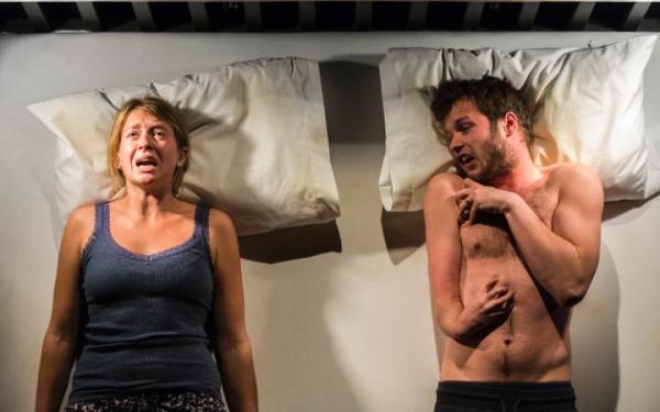 A young woman wearing a blue singlet lying on a bed with an anxious expression on her face. Next to her is a young man also lying on his back. He is only wearing black shorts.