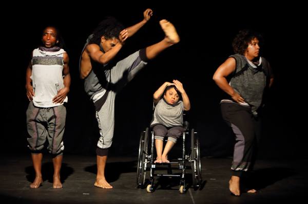 From left to right, two male dancers, one standing facing audience, the second lifting his arms and one leg high over the third dancer who is seated in a wheelchair. She leans away from him and holds her hands on her head. The last dancer, a woman, stands facing the away from the other dancers. They are all dressed in casual pants and tee shirts in various shades of grey. 