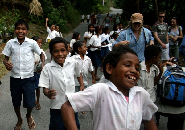Timorese school children dressed in uniform - white shirts and grey shorts and skirts walkingand running along a road surrounded by greenery. They are having fun. Three Australian college students in their midst.