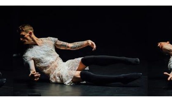 Colour Photograph of Dan Daw performing. He is dressed in a flimsy, short white lace dress and wears black thigh-length stockings. He is partially sitting on a black stage with black background, leaning back, head turned looking at the floor, arms stretched out slight to the front of his body. His legs are raised off the floor. He has short, fair hair and tattoos on his arms.