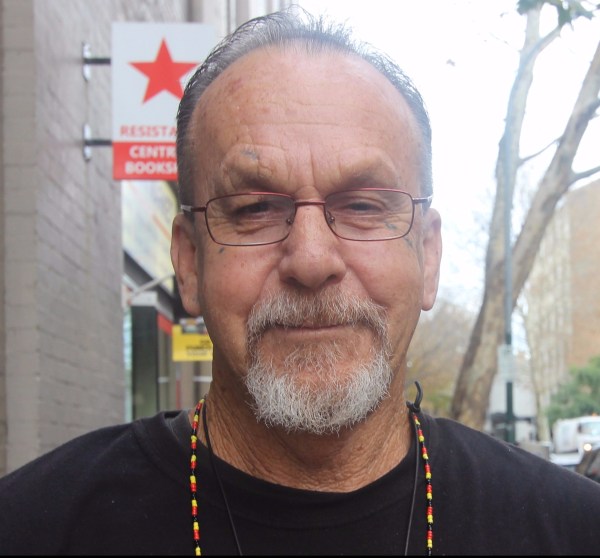 Head shot of Ken Canning. He is wearing glasses, and a long necklace of small, red, gold and black beads (Aboriginal colours). He has a greying mustache and beard, and he is smiling.