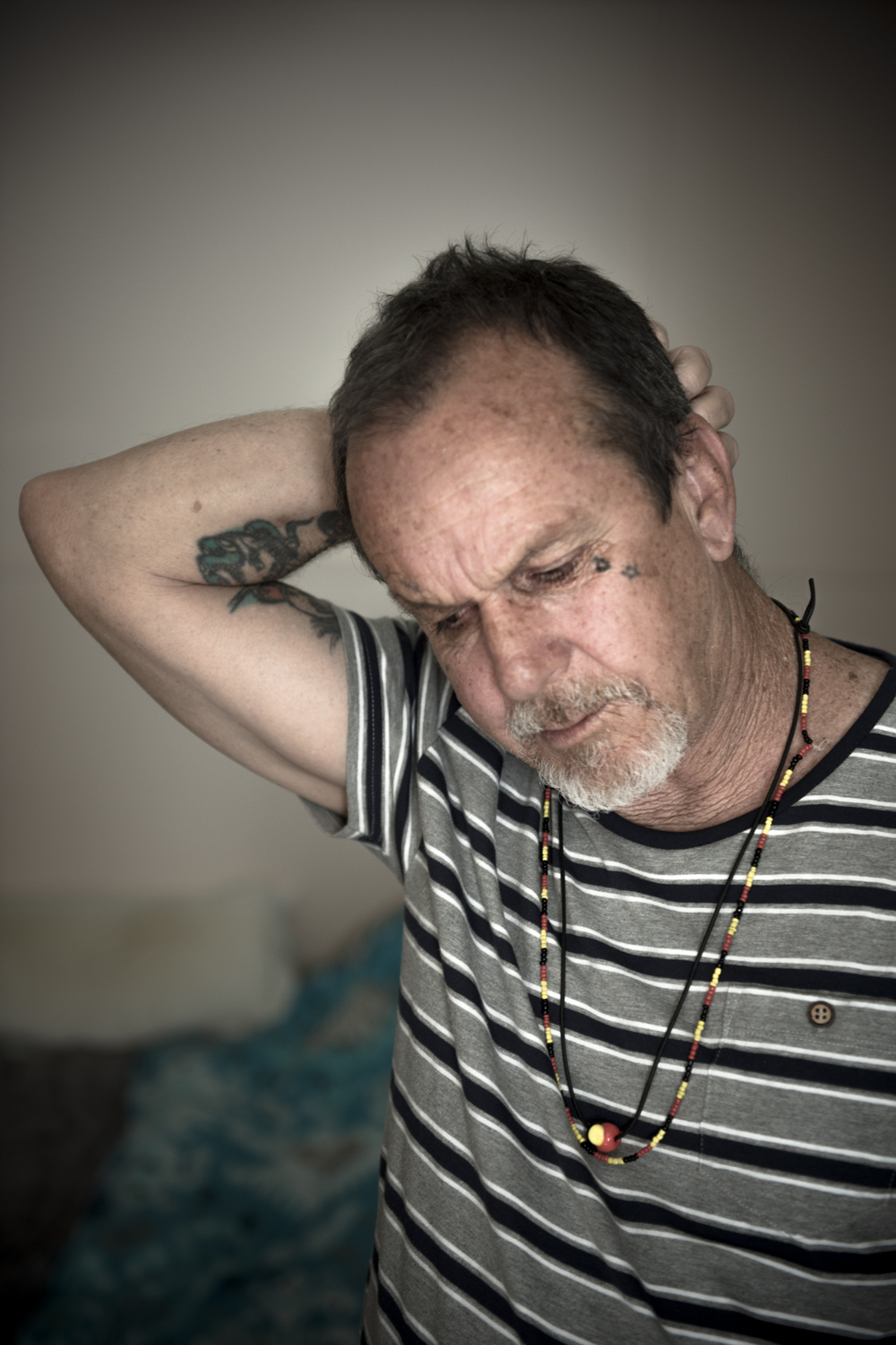 Colour photograph, interior, mid shot of Ken Canning standing in his apartment. Right hand clasped around the back of his head. He wears a striped grey, white and black T-shirt and two necklaces in the colours of the Aboriginal flag.