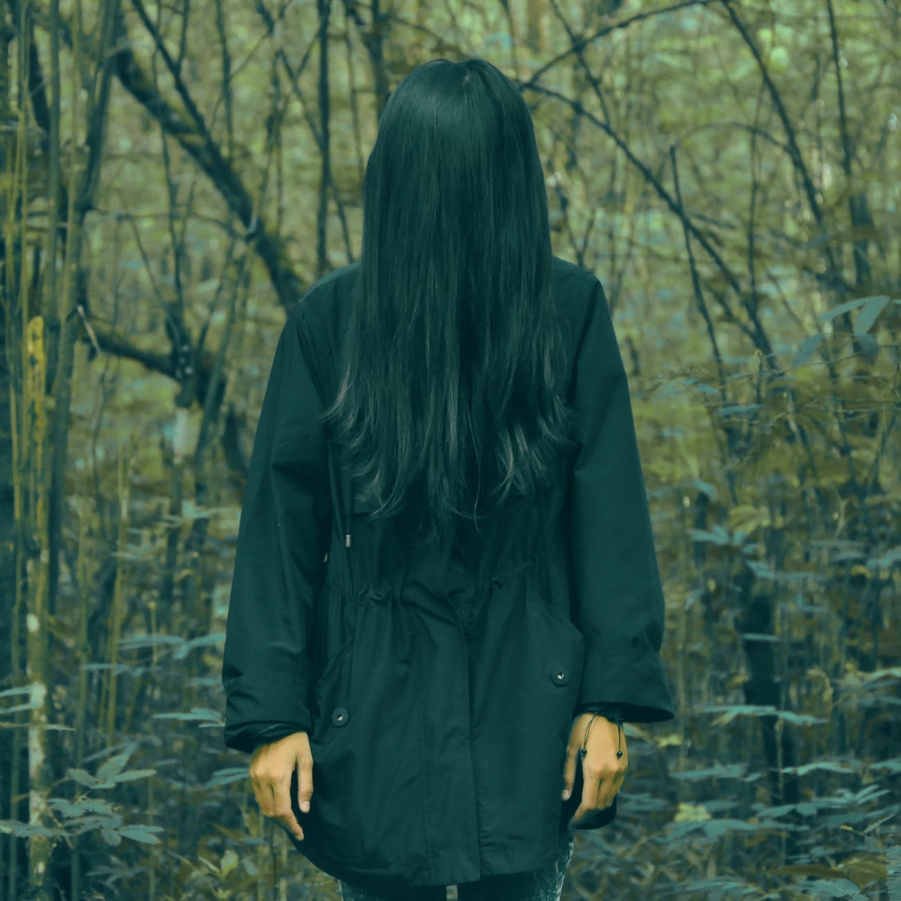 Colour photograph of a woman standing in a black coat in a forest. Her face is completely covered by her long black hair.