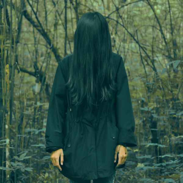 Colour photograph of a woman standing in a black coat in a forest. Her face is completely covered by her long black hair.