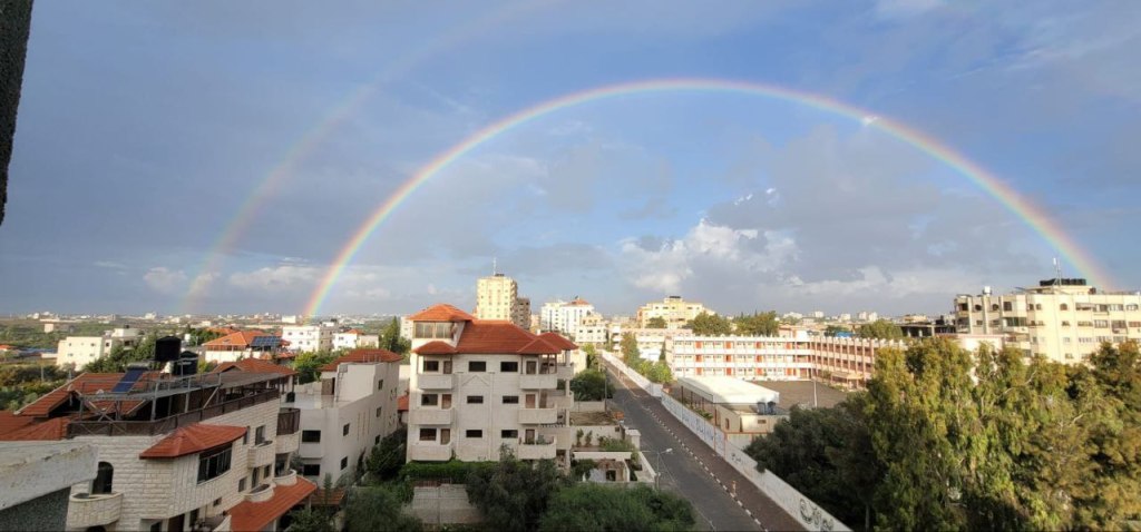 Al-Zahra showing white apartment buildings with red roofs, a blue sky and two rainbows stretching over the city